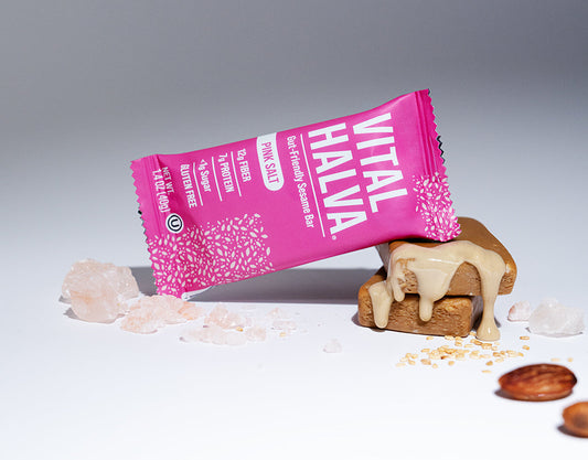 A pink wrapped sesame bar labeled 'VITAL HALVA' lies on its side on top of a sesame snack bar with beige tahini drizzle, surrounded by pink salt crystals and almonds on a white surface against a gray background.