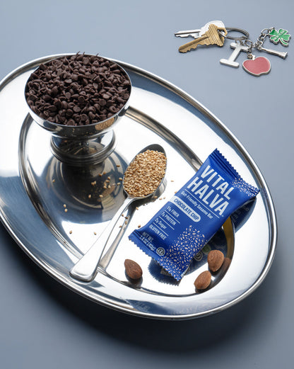 A silver oval tray on a gray background containing a bowl of dark chocolate chips, a spoon with sesame, a blue Vital Halva sesame snack bar, and a sesame seeds. A NYC keychain with a heart and tree charm is visible in the upper right.