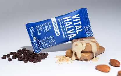 A Vital Halva chocolate chip sesame snack bar in its blue wrapper, shown lying on its side next to a tahini topped sesame snack bar, with chocolate chips, crumbs, and almonds scattered around on a light gray background.
