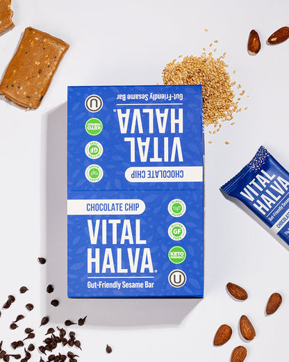 Vital Halva Chocolate Chip snack bar packaging shown on a clean white background with scattered chocolate pieces, almonds, and crumbs around it. The main box is blue with white text and green circular icons, and a smaller wrapped bar is placed to the right.