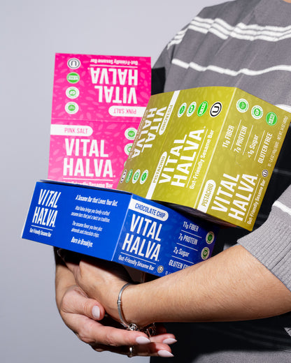 A person in a gray striped shirt holds three colorful Vital Halva sesame snack bar boxes stacked together against a plain light gray background. The boxes are pink, blue, and green with white text.