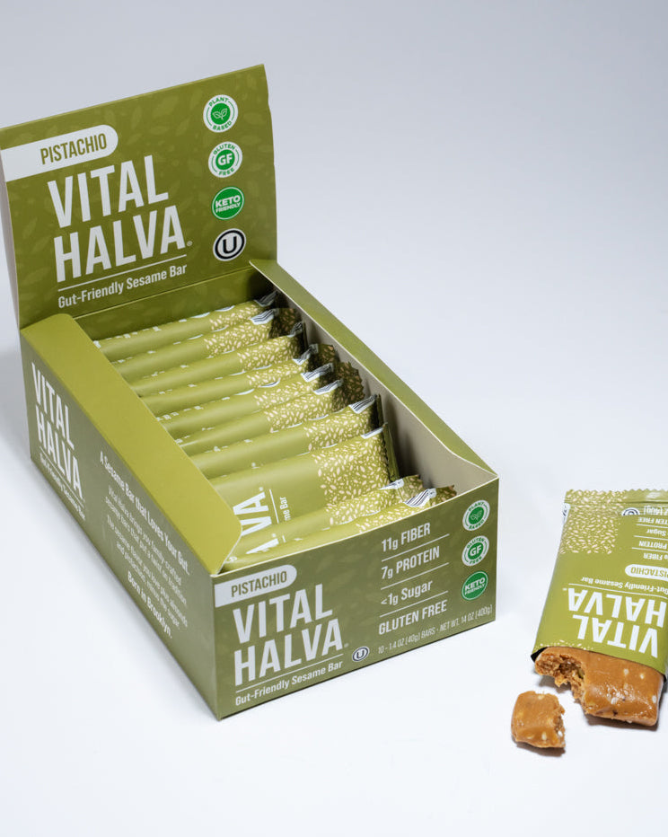 A box of Pistachio Vital Halva sesame snack bars with several individual wrapped bars and one loose bar placed beside it on a clean white studio background.