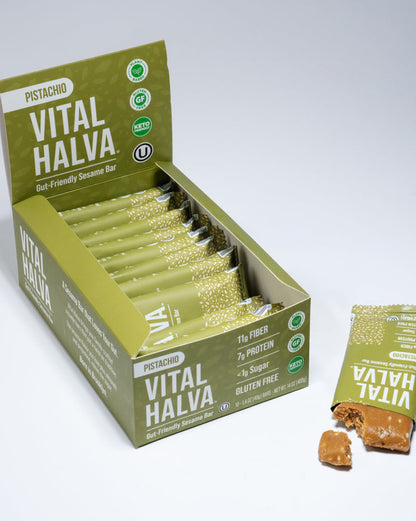 A box of Pistachio Vital Halva sesame snack bars with several individual wrapped bars and one loose bar placed beside it on a clean white studio background.