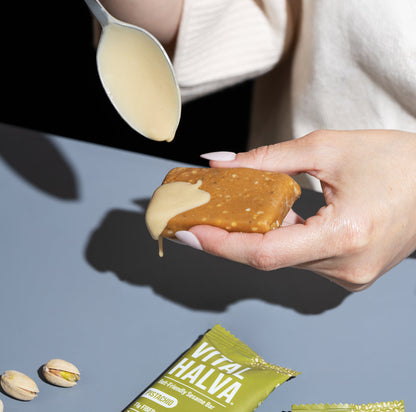 A person holds a piece of Vital Halva sesame snack bar Pistachio flavor, with tahini being drizzled over it from a spoon. Two green packets of Vital Halva and two pistachios are visible on a gray surface below.