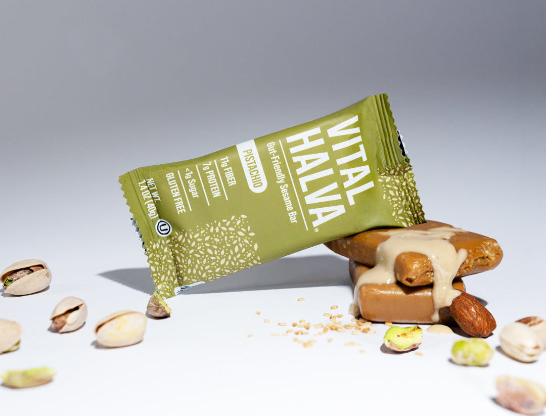 A Vital Halva sesame snack bar in its green wrapper, shown lying on its side next to a tahini-topped sesame snack bar with pistachios scattered on a white surface against a gray background.