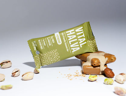 A Vital Halva sesame snack bar in its green wrapper, shown lying on its side next to a tahini-topped sesame snack bar with pistachios scattered on a white surface against a gray background.