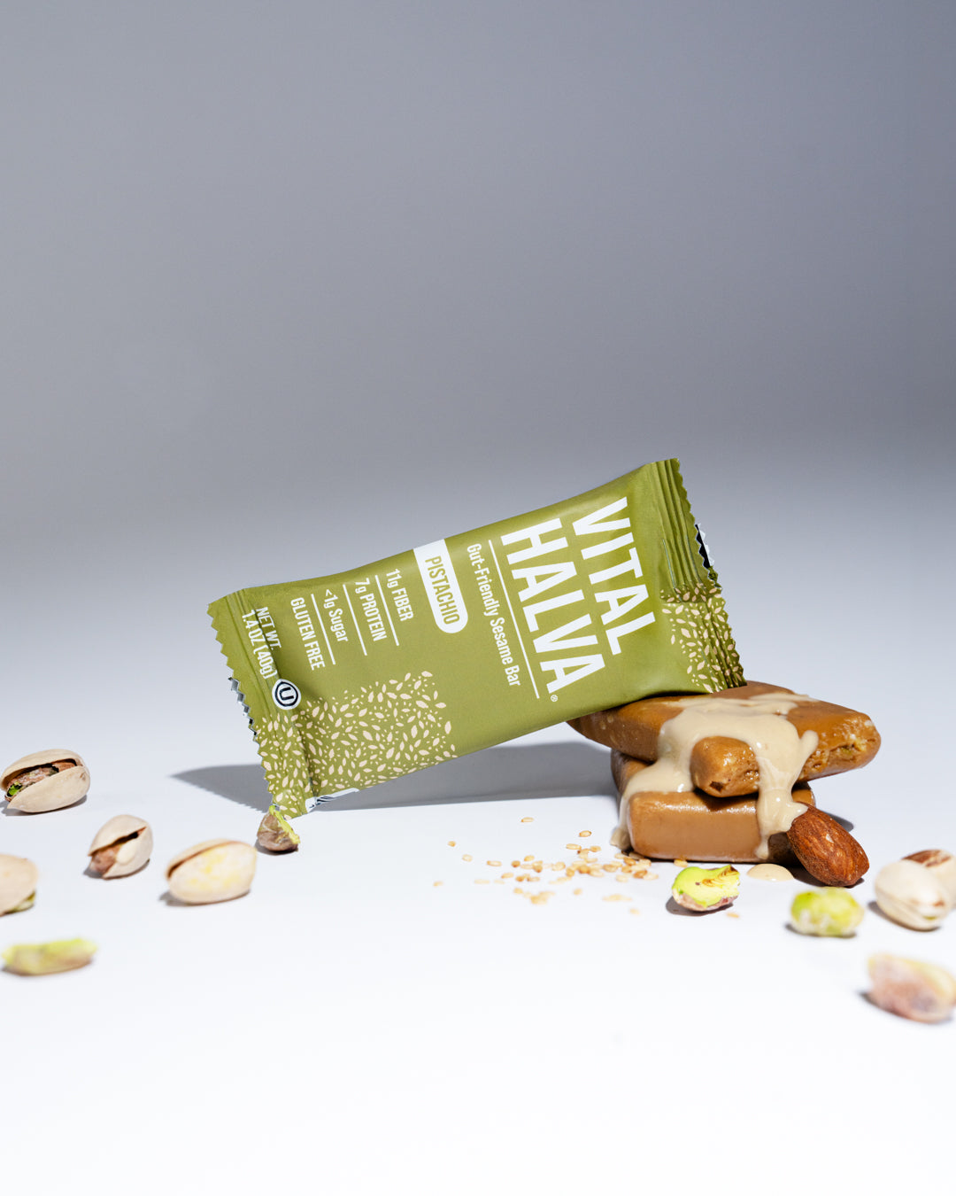 A Vital Halva sesame snack bar in its green wrapper, shown lying on its side next to a tahini-topped sesame snack bar with almonds and pistachios scattered on a white surface against a gray background.
