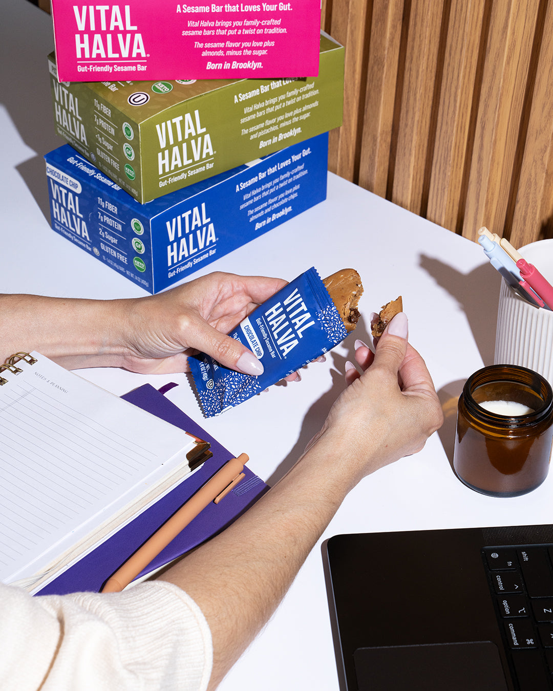 A person at a desk holds a blue Vital Halva snack bar, opened, with stacked Vital Halva product boxes in pink, green, and blue behind them on a white desk.